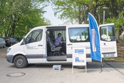 Weißer Sprinter seitlich zu sehen mit offener Schiebetür. Innen eine Gesprächsleiterin von Sprachbrücke-Hamburg e. V. sitzend. Davor ein Plakataufsteller und ein Werbesegel