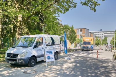 Weißer Sprinter seitlich zu sehen mit offener Schiebetür. Innen eine Gesprächsleiterin von Sprachbrücke-Hamburg e. V. sitzend. Davor ein Plakataufsteller und ein Werbesegel. Im Hintergrund ein Gebäude