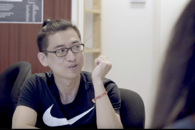 Ein junger Mann asiatischer Herkunft mit Brille, Kurzarm-Shirt und zurückgebundenen Haaren sitzt an einem Tisch und schaut zur Seite im Gespräch mit einer oder mehreren Personen