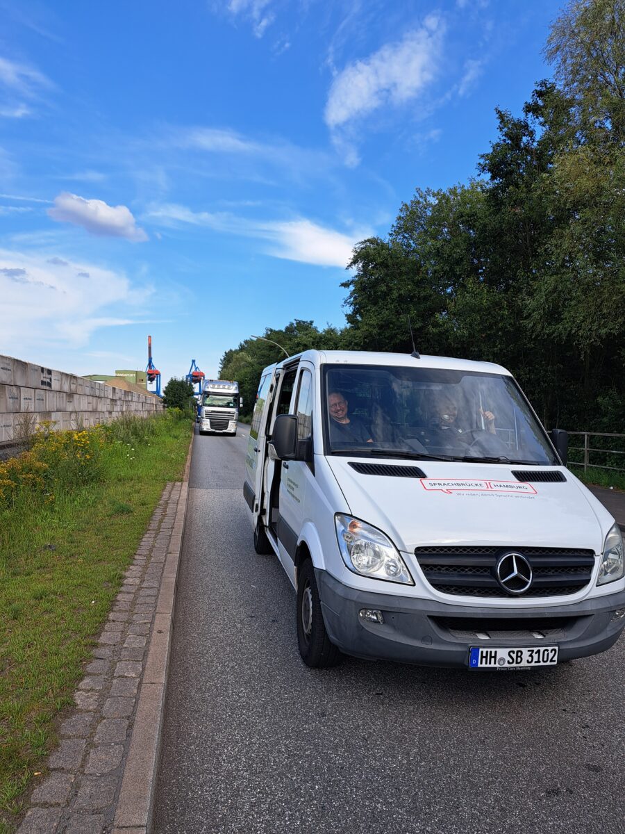 Ein weißer Transpoerter steht mit 2 Fahrern in einer langen Einfahrt. Daneben ist eine kleine Grünfläche. Dahinter fährt ein Lkw ein.