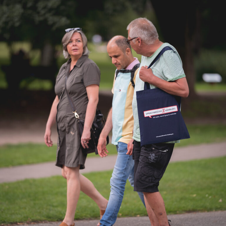 Eine Frau und zwei Männer während einer Unterwegsgesprächsrunde von Sprachbrücke-Hamburg e.V. spazieren. Im Hintergrund ist eine Hecke oder Bäume zu sehen.