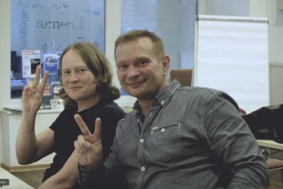 Eine Frau und ein Mann während einer Gesprächsrunde von Sprachbrücke-Hamburg e.V. lächeln in die Kamera und zeigen das Victory-Symbol mit ihrer jeweils linken Hand.